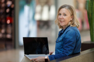 Blonde Frau sitzt an ihrem Laptop und schaut über die Schulter.