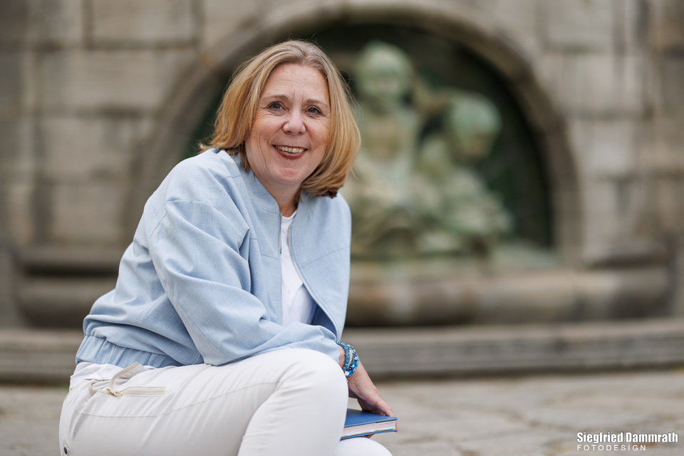 Blonde Frau vor einem Brunnen lächelt in die Kamera.